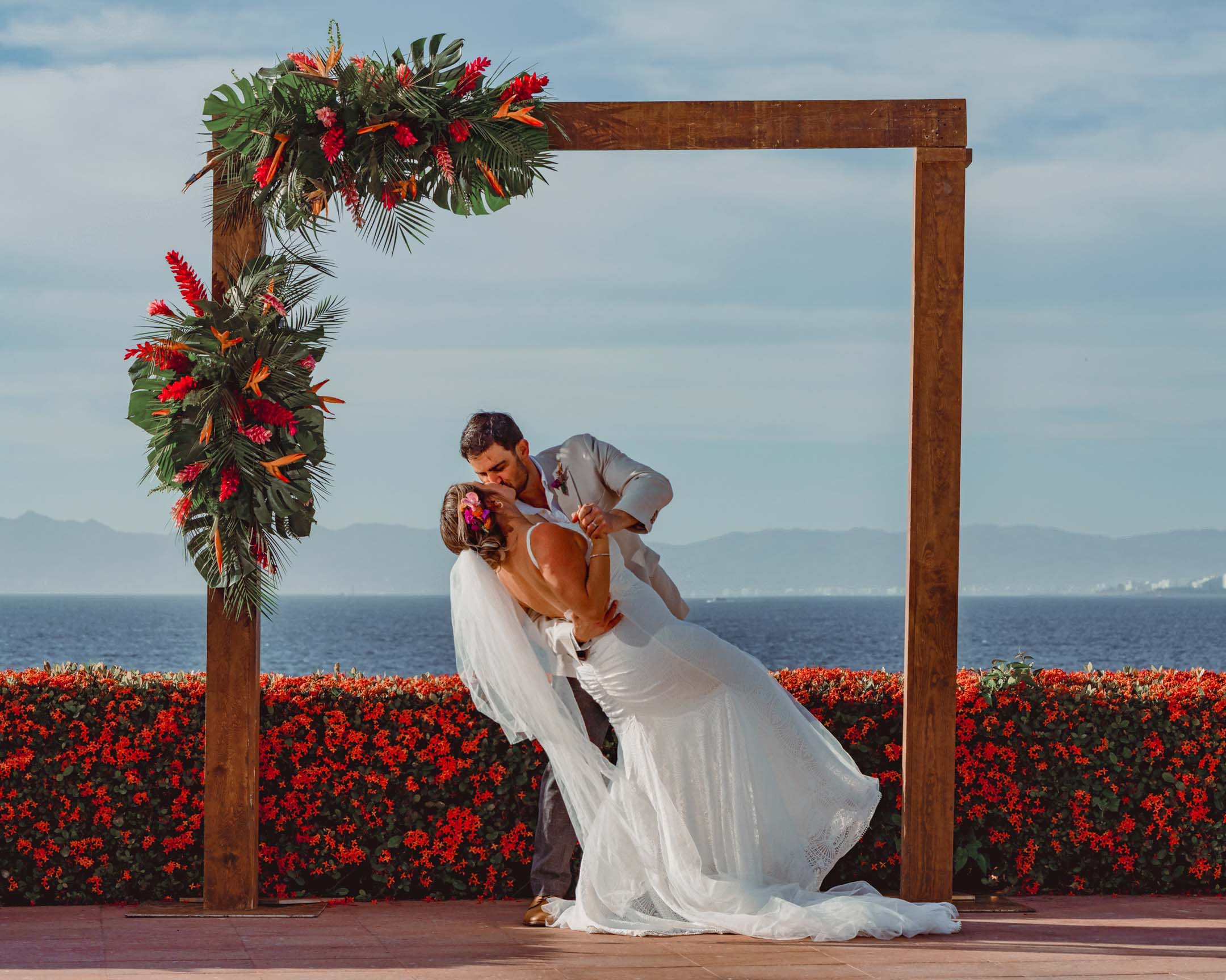 Bride and groom kiss under a tropical floral arch overlooking the bay